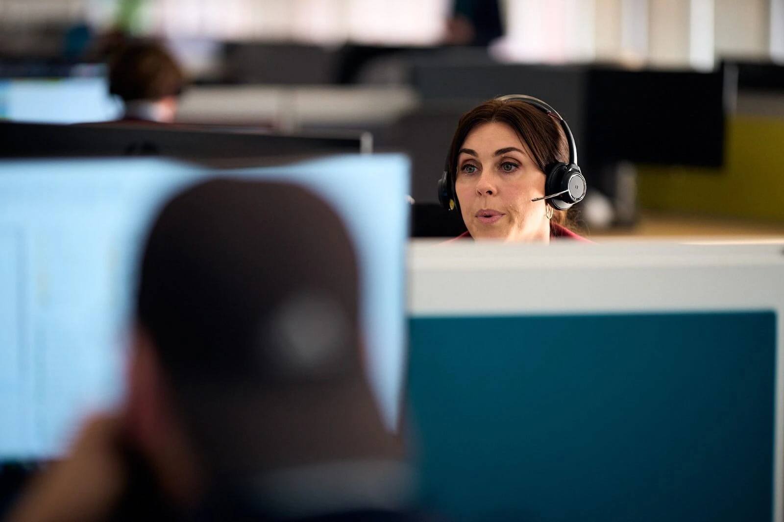 Woman wearing a headset in a shared office environment, focusing on her computer screen while other desks and monitors are blurred in the background.