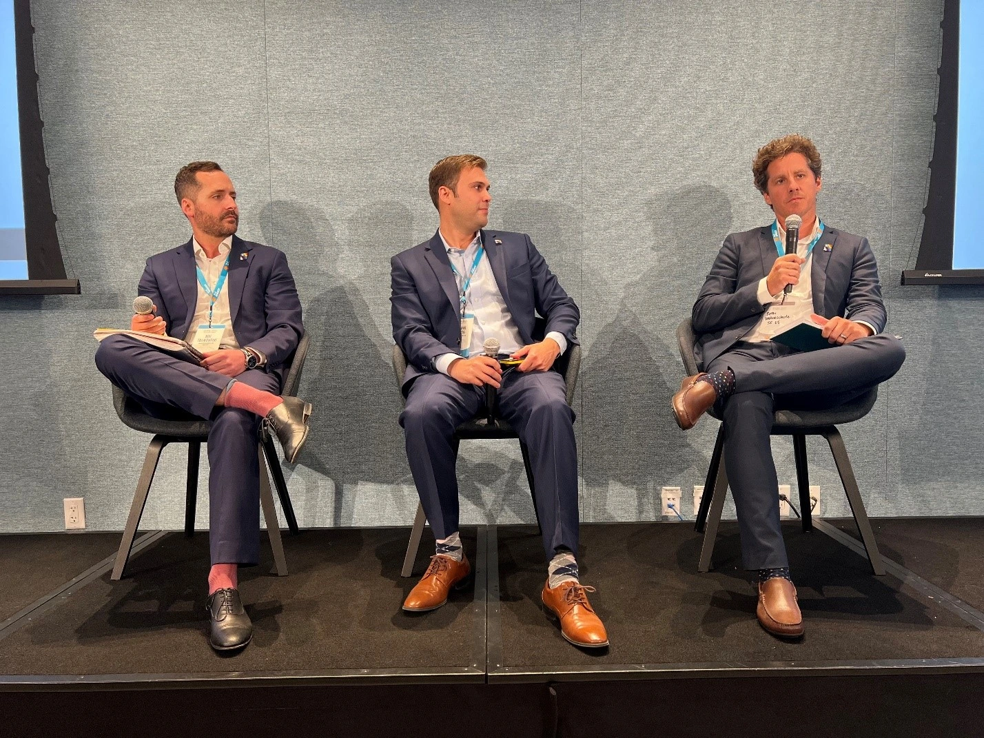 Three men in suits seated on stage with microphones at a panel discussion.