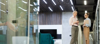 Two professionals in a modern office hallway having a conversation, one holding a coffee cup and the other holding a clipboard, with office furniture and reflections visible in the background.